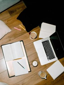 Top view of a creative workspace with laptop, notebooks, and coffee on a wooden floor.